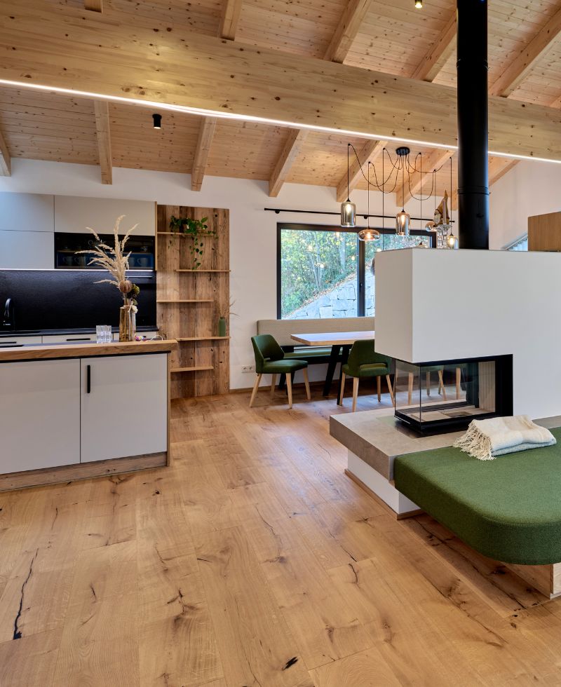 Modern eingerichteter Wohnraum mit offener Küche, Essbereich und Kamin. Der Raum hat eine hohe Holzdecke mit sichtbaren Balken, hellen Holzboden und große Fenster, die viel Tageslicht hereinlassen. Die Küche ist in Grau und Holz gestaltet, der Essbereich verfügt über grüne Stühle, und neben dem Kamin befindet sich eine grüne gepolsterte Sitzbank.
